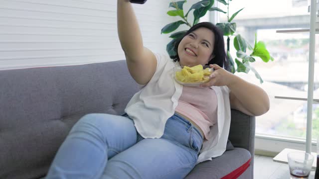 Cheerful Asian chubby woman smiling on the sofa, holding potato chips, taking selfie on smartphone.