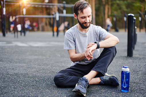 Young man sitting on the ground in the park getting ready for his outdoor workout, setting up smart watch mobile tracking application