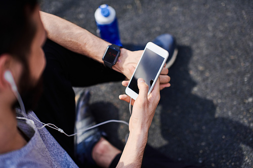 Close up of a man sitting on the ground in the park getting ready for his outdoor workout, setting up smart phone and watch mobile tracking application, top view, listening to music