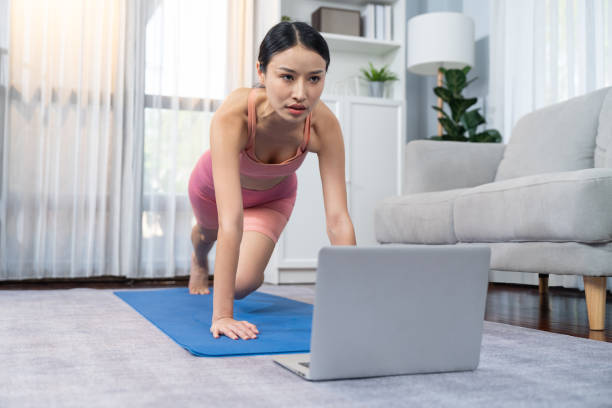 Asian woman in sportswear doing burpee on with online exercise video. Vigorous Asian woman in sportswear doing burpee on exercising mat as home workout training routine. Attractive girl engage in her pursuit of healthy lifestyle with online exercise training video. Vigorous burpee stock pictures, royalty-free photos & images