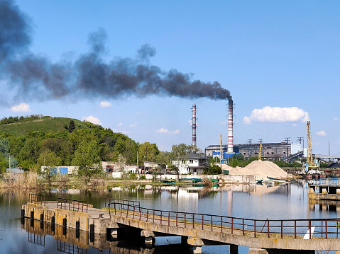 In the background, black smoke is billowing from the plant's pipes against a blue sky. The river is in the foreground. Environmental pollution