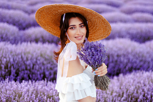 A young girl,beautiful woman portrait in lavender field in white dress and straw hat holding a bouquet of lavender flowers.Front view of beautiful girl in a white dress and hat in a lavender field at sunset