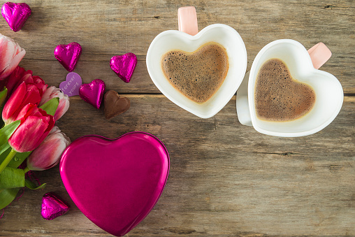 Two heart shaped cups of coffee, heart shaped purple gift box with chocolates and bouquet of red tulips on a old wooden background; top view, copy space