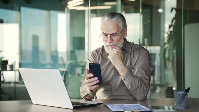 Frustrated stressed senior gray haired bearded businessman reads bad news on phone sitting at workplace in business office.