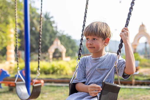 Portrait of boy in gray T-shirt and shorts who is sitting alone on swing on playground on summer day. Summer holidays. Children's active outdoor games. Children's problems, no friends.