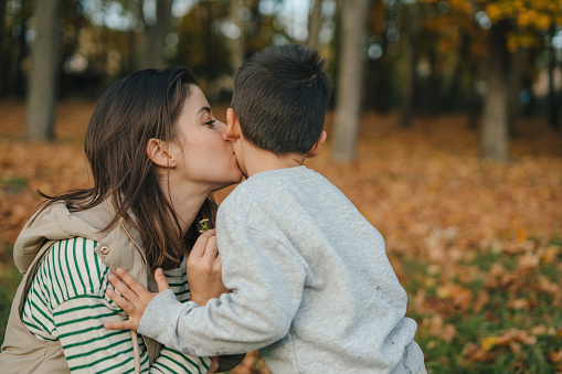 Mother kissing child on cheek, having a walk in autumn forest in nature. Family holiday, spending time together at sunset.