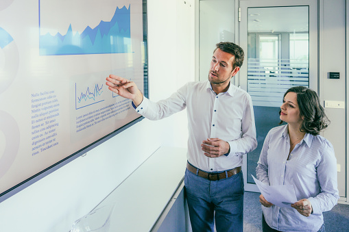 Businesswoman and businessman discussing finance in front of a screen with a graph.