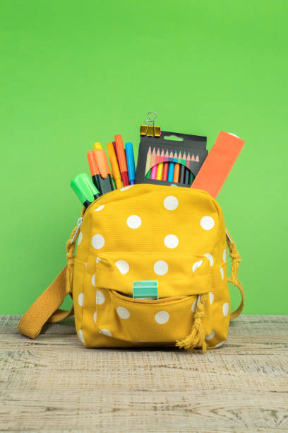 istock Yellow backpack with white polka dots with different colorful stationery on table. Bright green background. Back to school. 1982309679