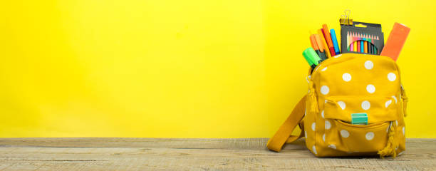istock Yellow backpack with white polka dots with different colorful stationery on table. Yellow background. Back to school. Banner design. 1982322381