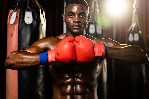 Boxing fighter posing, African American Black boxer put his hand or fist wearing glove together in front in aggressive stance and ready to fight at gym with kicking bag and boxing equipment. Impetus