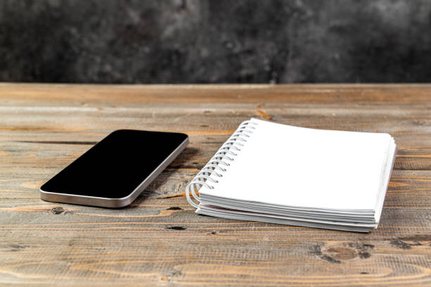 Closeup view of mobile phone with blank screen and an empty notebook on a wooden background. Blank mobile phone screen and empty notebook for copy space