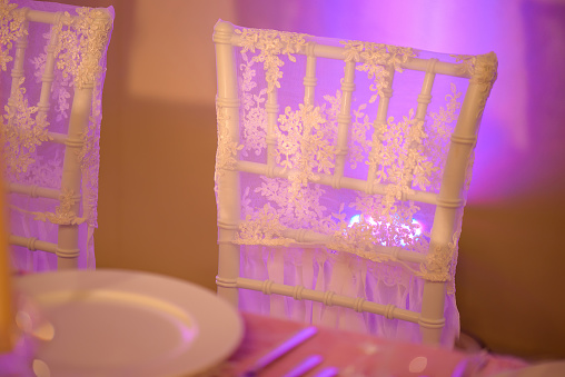 Table setup for a formal dining experience such a wedding or a banquet with focus on a white chair backrest covered in a thin, see-through silky cloth with a purple light delicately reflecting through.
