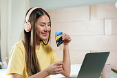 Young woman in yellow top presents credit card, ready for online transaction on laptop. Confidently holding a credit card, a young adult female smiles as she prepares to shop online with laptop.