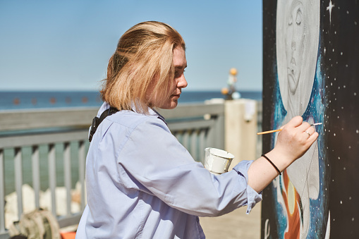 Young female painter passionately paints picture with paintbrush for outdoor street exhibition, female artist engrossed in creating vibrant artwork at bright sunny day