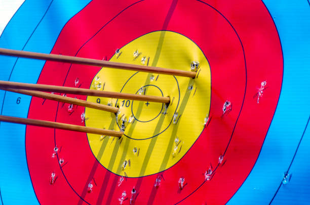 A teenage Caucasian female archer in a studio setting aims her bow and arrow with precision. Wearing casual athletic attire, she demonstrates concentration and skill against a dark background, highlighting the target with vibrant colors.