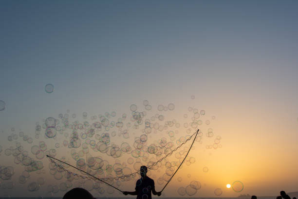 A man in silhouette makes lots of soap bubbles with a soap bubble stick, they float in the orange sky at sunset