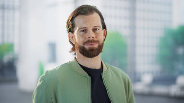 istock Handsome Young Man with a Beard Standing in a Modern Office Building Space. Young Happy Creative Caucasian Male Looking at Camera, Gently Smiling. Portrait of a Casually Dressed Man at Daytime 1982622952
