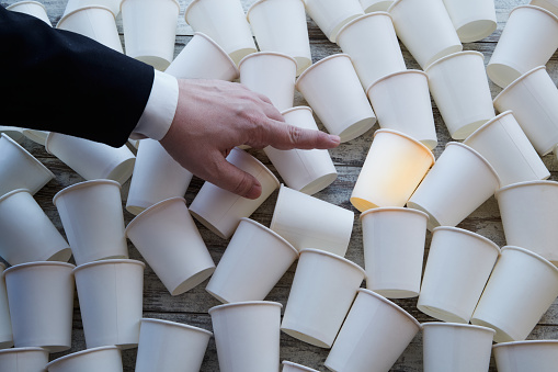 White paper cups scattered on the table and a businessmanâs hand pointing to the brightest one. The problem of choice, find a solution option from many