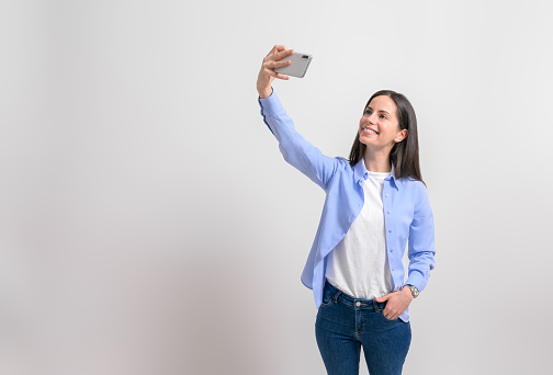 Smiling beautiful entrepreneur with hand in pocket taking selfie over cellphone on white background