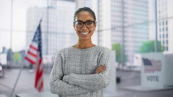 Portrait of a Beautiful African American Female Posing with Big Bright Smile with American Flag in the Background. Successful Young Black Woman Looking at Camera, Standing with Crossed Arms