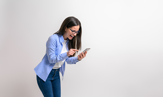 Young businesswoman laughing and texting online over smart phone while standing on white background