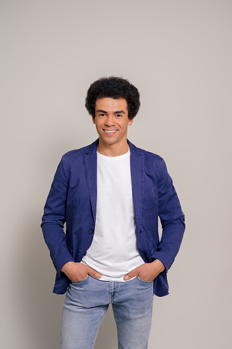 Portrait of happy male professional with afro hair and hands in pockets posing over white background