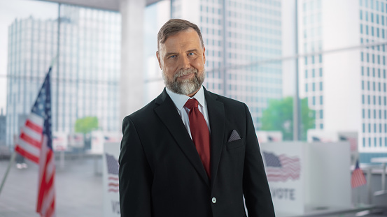 Portrait of an Adult Male in a Suit, United States Elections Candidate, Posing for Camera, Smiling. Caucasian Senior Man Standing in a Modern Polling Station with American Flag in the Background