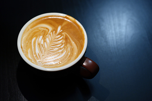 In a close-up shot, a hot latte is presented in a brown coffee cup with latte art, placed on a black wood table at a cafe. This image represents the concept of food and drink, capturing the aesthetic appeal of a freshly prepared beverage.