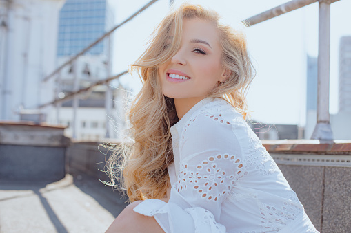 Positive emotions. Enjoying life and summer day is demonstrated by charming blonde in white dress with long luxurious hair, posing on roof ofcity building. Freedom and independence. Confidence .
