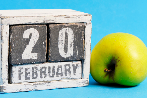 February 20th on wooden calendar and green apple on blue background. Apple Day USA