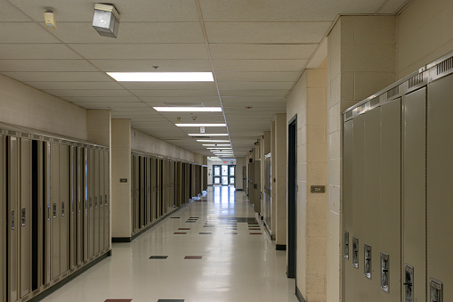 Empty school hallway - lined with closed lockers - beige walls - tiled floor - ceiling lights illuminating space - exit doors at the end. Taken in Toronto, Canada.