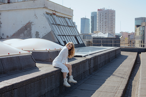Young blonde woman in white dress and high white shoes is sitting on roof of building against backdrop of high-rise buildings. Beautiful women. Urban style. Modern free women. Rhythms of the city.