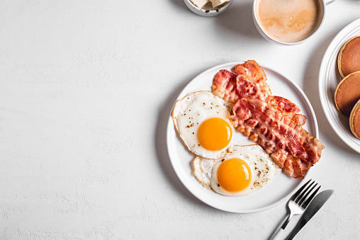 Breakfast with fried eggs, bacon, pancakes and coffee, top view, banner. Fried sunny side eggs with bacon on plate and morning cappuccino coffee. American breakfast.