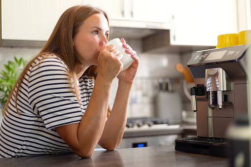 A thoughtful woman savoring a hot drink in a mug, sitting in a well-equipped, contemporary kitchen with a coffee machine.