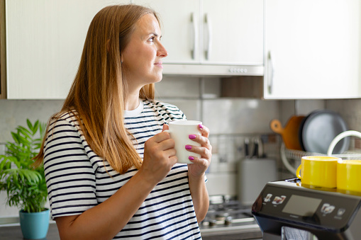 Contemplative woman holding a coffee mug in a sunlit kitchen.