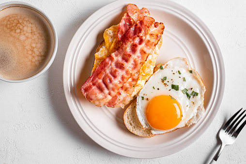 Breakfast sandwiches with fried egg, bacon, scrumbled egg and coffee on white table, top view. Fried eggs with bacon on toasts and morning cappuccino coffee.