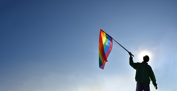 Rainbow flag raising, LGBT simbol, holding in hands of boy against clear bluesky background, soft and  selective focus, concept for LGBT celebration in pride month, June, around the world, copy space.