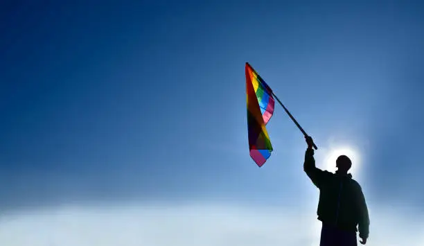 Rainbow flag raising, LGBT simbol, holding in hands of boy against clear bluesky background, soft and  selective focus, concept for LGBT celebration in pride month, June, around the world, copy space.