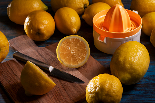 Cutting board with a knife and a lemon freshly cut in half, surrounded by lemons and a manual juicer.