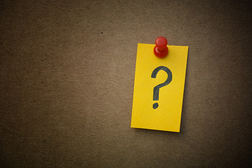 A yellow paper note with a question mark on it pinned to a cardboard background. Closeup.