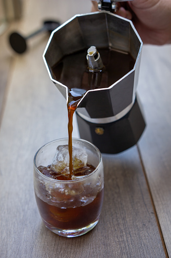 a hand pouring coffee from a moka pot into a glass with ice.