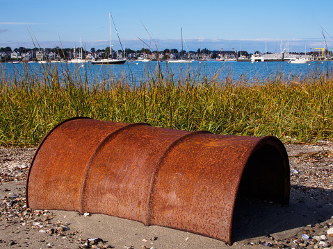 A detailed capture unveils the rustic beauty of a half-buried, weathered barrel in the sandy embrace of Coughlin Park, Winthrop, just outside Boston, Massachusetts, USA. Bathed in sunlight, the scene unfolds against a backdrop of shimmering water and sailboats, weaving a tale of coastal allure and time's gentle touch.