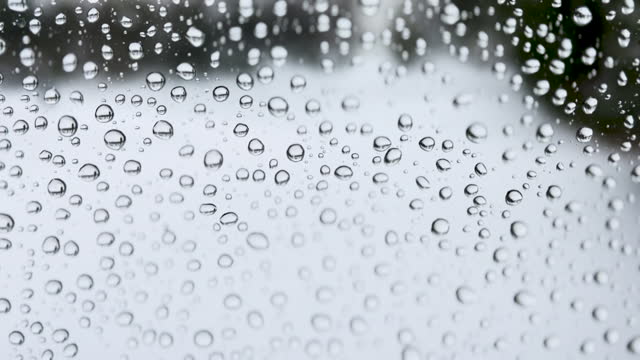 Close-up of water drops on glass, Extreme macro video closeup of rain drops collecting on a transparent glass window pane on an overcast, gloomy, cloudy, rainy weather day with focus on the droplets as they wiggle in the wind