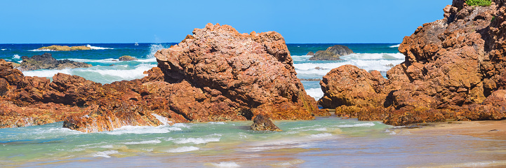 Australian coast with volcanic rocks on the shore, view from the beach to the horizon with blue water and waves breaking on the rocks, summer sunny day.