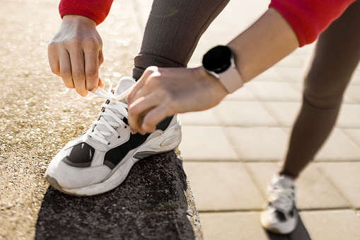 Detail of a woman's hands lacing up her running shoes, capturing the initial step in her fitness regimen against an outdoor backdrop.