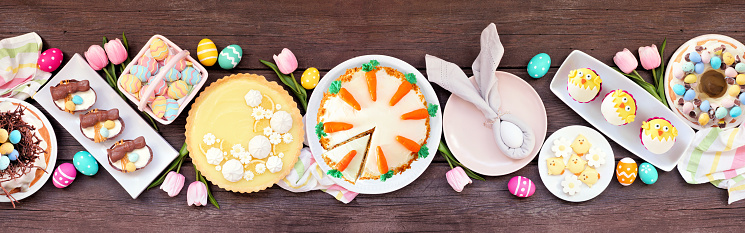 Easter or spring dessert food table scene. Above view over a dark wood banner background. Lemon tart, cupcakes, Easter egg and carrot cakes and various sweets.