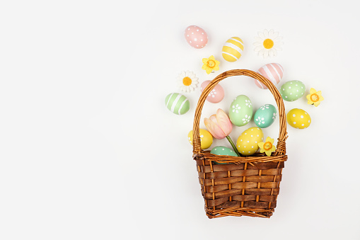 Spilling Easter basket of colorful pastel hand painted Easter Eggs and flowers. Top down view over a white background.