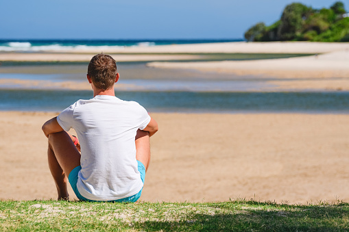 Young man dressed in sportswear sits by the beach and looks towards the sea at low tide, seaside landscape on a summer sunny day.