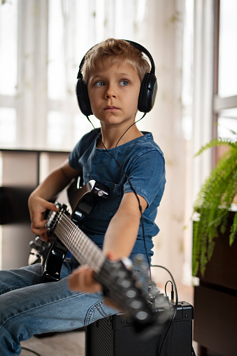 Little boy learning to play electric guitar. The boy is wearing a blue jeans t-shirt and blue jeans trousers. He is sitting on a large black amp and he is very focused on strumming the black guitar.\nShot with Nikon D800