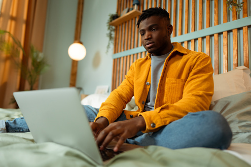 Portrait of handsome African American professional programmer sitting on bed, using laptop, at home, copywriter typing on keyboard. Concept of technology, remote job